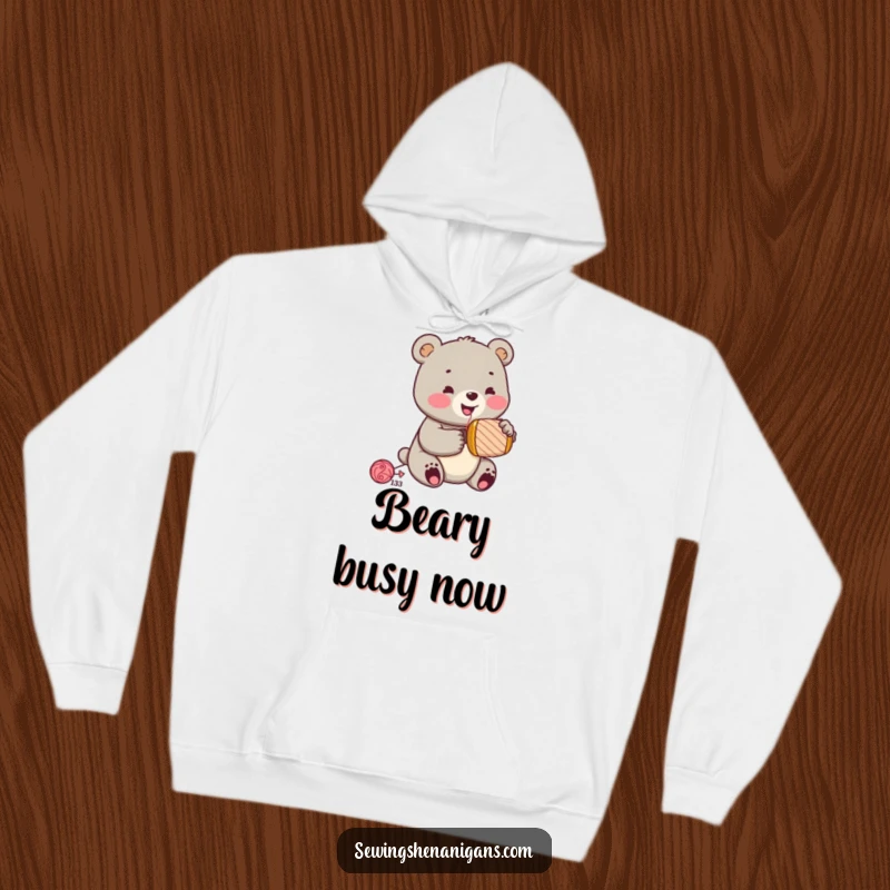 Funny jolly bear cub wearing a sewing thimble, enthusiastically hammering a spool, on a cozy hoodie, a warm and funny gift.